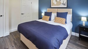 A neatly made bed with a blue and beige bedspread, two bedside tables with lamps, and a framed artwork above the bed. - Host & Stay | Apartment 3, Upton House (Northampton)