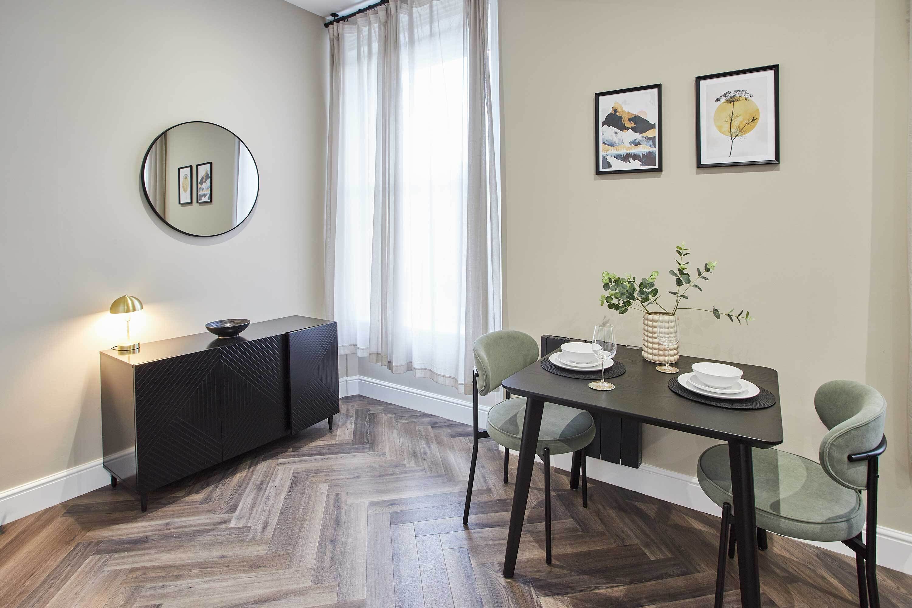 A dining area with a herringbone wood floor, a black dining table, four chairs, a black console, and a round mirror on the wall.
