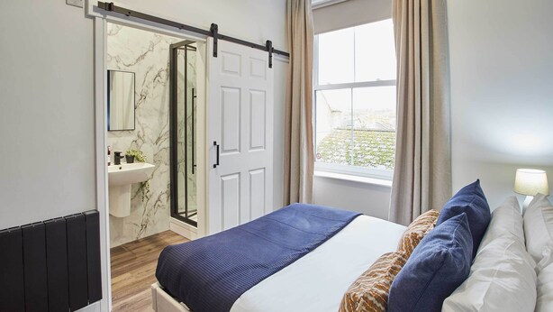 A bedroom with a bed, a window with curtains, a door to a bathroom, and a radiator. - Host & Stay | Apartment 4, Upton House (Northampton)