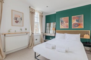 A hotel room with a large bed, a window with curtains, a radiator, a wooden shelf with plants, and artwork on the walls. - Perfect 2BD Duplex with Private Balcony, Islington (Greater London)