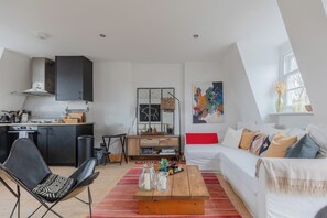 A modern living room with a white sofa, a wooden coffee table, and a black chair. There is a kitchen area with a stove and cabinets. - Perfect 2BD Duplex with Private Balcony, Islington (Greater London)