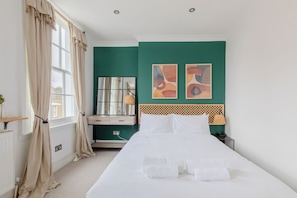Un lit bien fait, avec du linge de lit et des serviettes blanches, une table de chevet en bois avec une lampe, un mur d’accent vert et une fenêtre avec des rideaux.