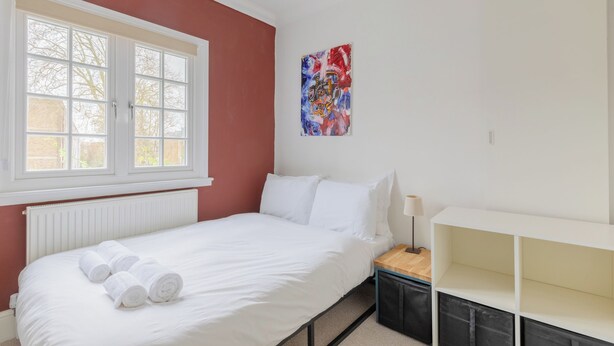 A bedroom with a bed, white bedding, a window with blinds, a radiator, a painting on the wall, and a bedside table with a lamp. - Perfect 2BD Duplex with Private Balcony, Islington (Greater London)