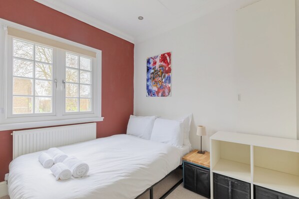 A bedroom with a bed, white bedding, a window with blinds, a radiator, a painting on the wall, and a bedside table with a lamp.