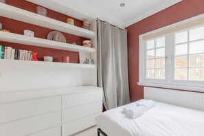 A bedroom with a white bed, a window with white shutters, a radiator, and a shelf with books and decorative items.