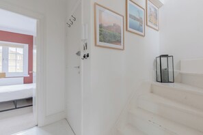 A white room with a bed, a door, and a staircase. There are framed pictures on the wall.