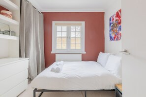 Une chambre à coucher avec un lit, une fenêtre, un radiateur, une commode blanche et une peinture abstraite aux couleurs vives accrochée au mur.