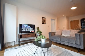 A modern living room with a flat-screen TV, a grey sofa, a coffee table, and a potted plant. - Suite Life 3: 2-Bed, 2-Bath in New Cross (Greater Manchester)