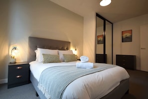 Un lit bien fait, avec une tête de lit grise, deux tables de chevet dotées de lampes et un grand miroir accroché au mur.