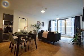 Une cuisine moderne équipée d’un four à micro-ondes, d’un four traditionnel, d’une table à manger avec des chaises, d’un canapé noir et d’une grande fenêtre offrant une vue sur l’immeuble.