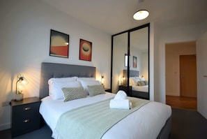 Ein ordentlich bezogenes Bett mit Kissen, ein Nachttisch mit Lampe und ein großer Spiegel.