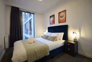 A hotel room with a bed, a nightstand, a window with curtains, and two wall art pieces. - Suite Life: 2-Bed, 1-Bath in New Cross (Greater Manchester)