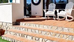Een modern terras buiten met witte schommelstoelen, een stenen trap en een groot raam met uitzicht op de omgeving.