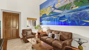 Un salon avec un canapé en cuir marron, une table basse en bois et un tableau représentant un paysage marin avec des bateaux et des poissons.