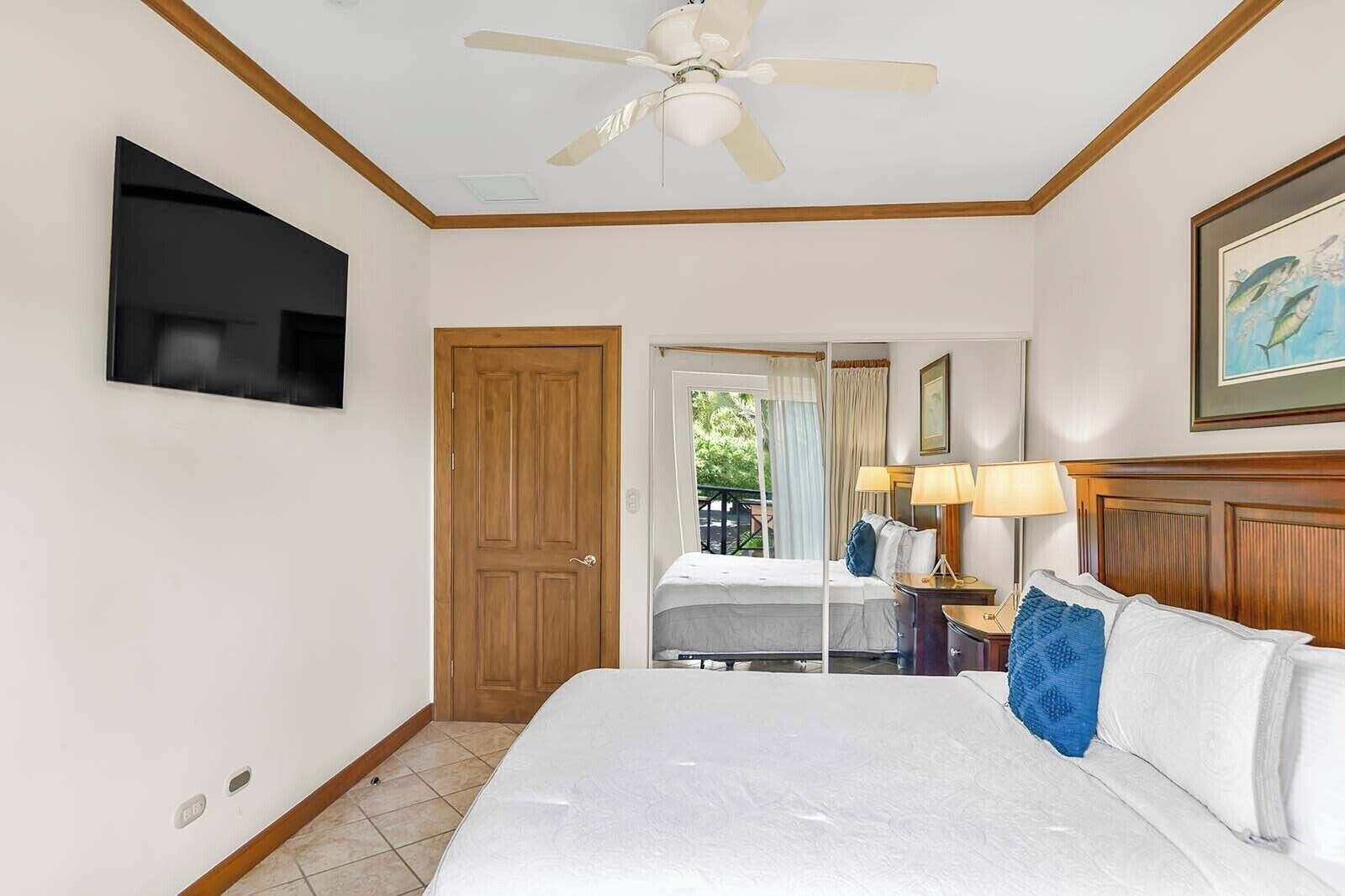 A hotel room with a bed, a ceiling fan, a TV mounted on the wall, a wooden door, and a window with curtains.