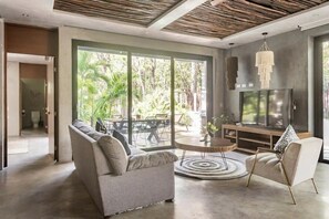 Ein modernes Wohnzimmer mit Sofa, Sessel und Fernseher, großen Fenstern mit Blick auf Grünflächen und einer Holzbalkendecke.