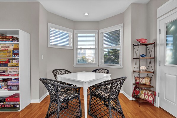 A dining area with a white table and black wicker chairs, a bookshelf with board games, and a metal shelf with decorative items.