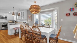 A modern kitchen with white cabinets, a central island, and a dining area with wicker chairs and a white table. - Seasaw Cottage - 5 bedroom pet friendly home on Wrightsville Beach (Wrightsville Beach)