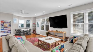 A living room with a sofa, coffee table, and TV. There is a bookshelf, a dining table with chairs, and a window with blinds. - Seasaw Cottage - 5 bedroom pet friendly home on Wrightsville Beach (Wrightsville Beach)