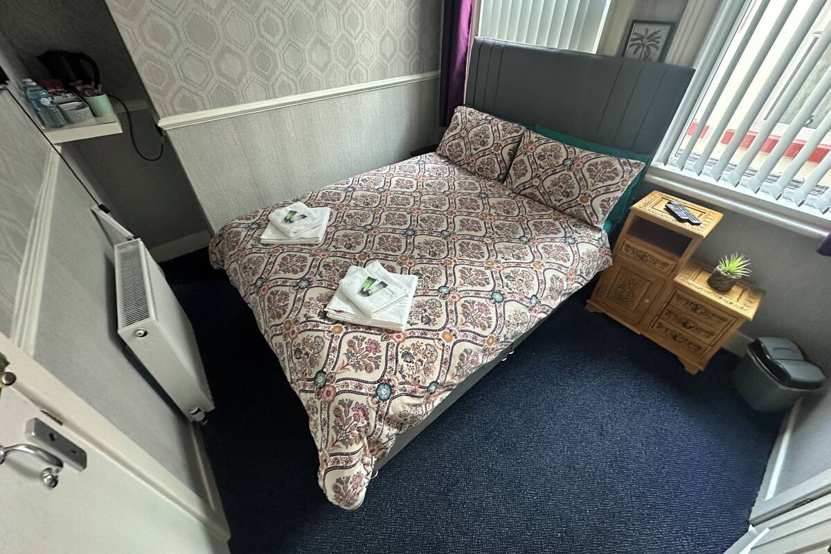 A neatly made bed with patterned bedding, a bedside table with a plant, and a window with blinds.