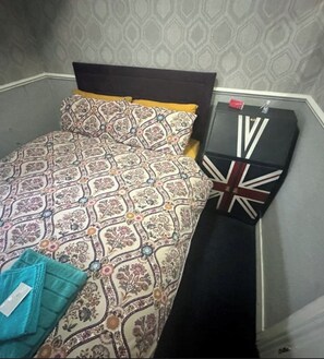 A small, neatly made bed with a patterned bedspread and pillows, a black suitcase with a Union Jack design, and a pair of turquoise slippers on the floor. - Runway Guest House - Adult Only (Blackpool)