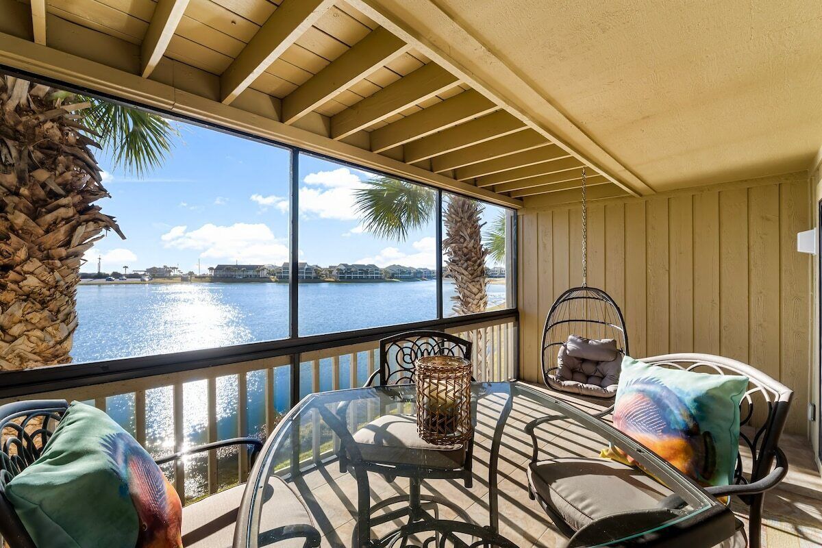 A covered outdoor seating area with a glass table and chairs, overlooking a body of water and palm trees.