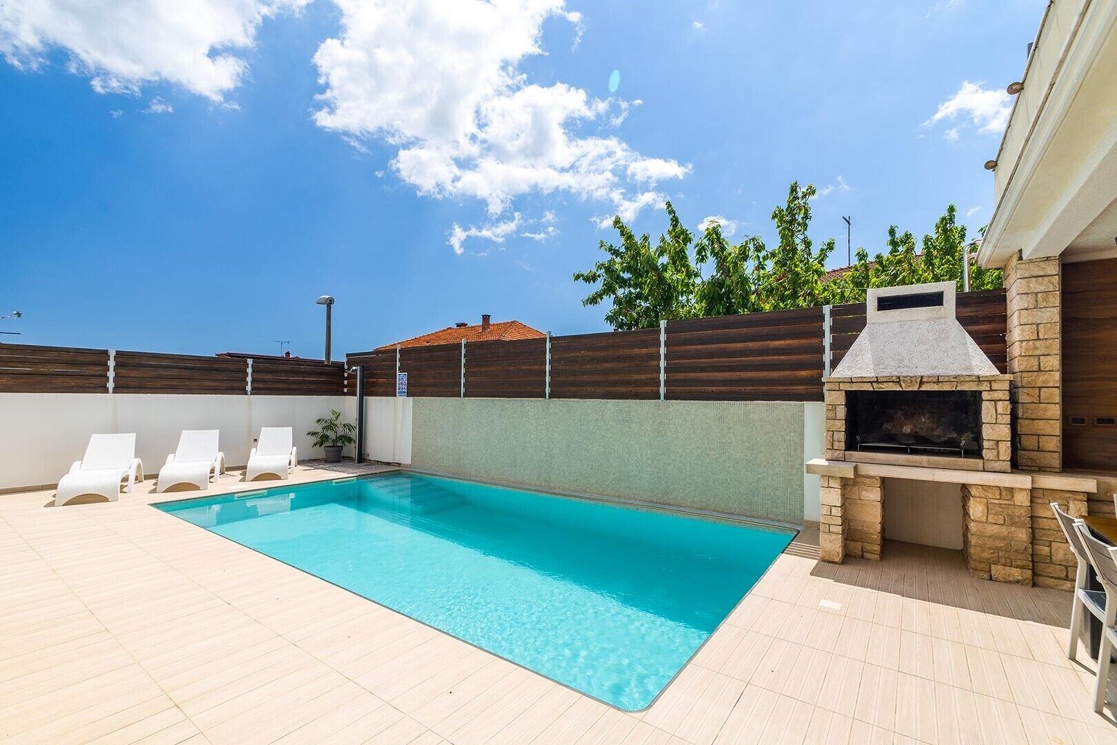 A swimming pool with lounge chairs, a barbecue area, and a wooden fence.