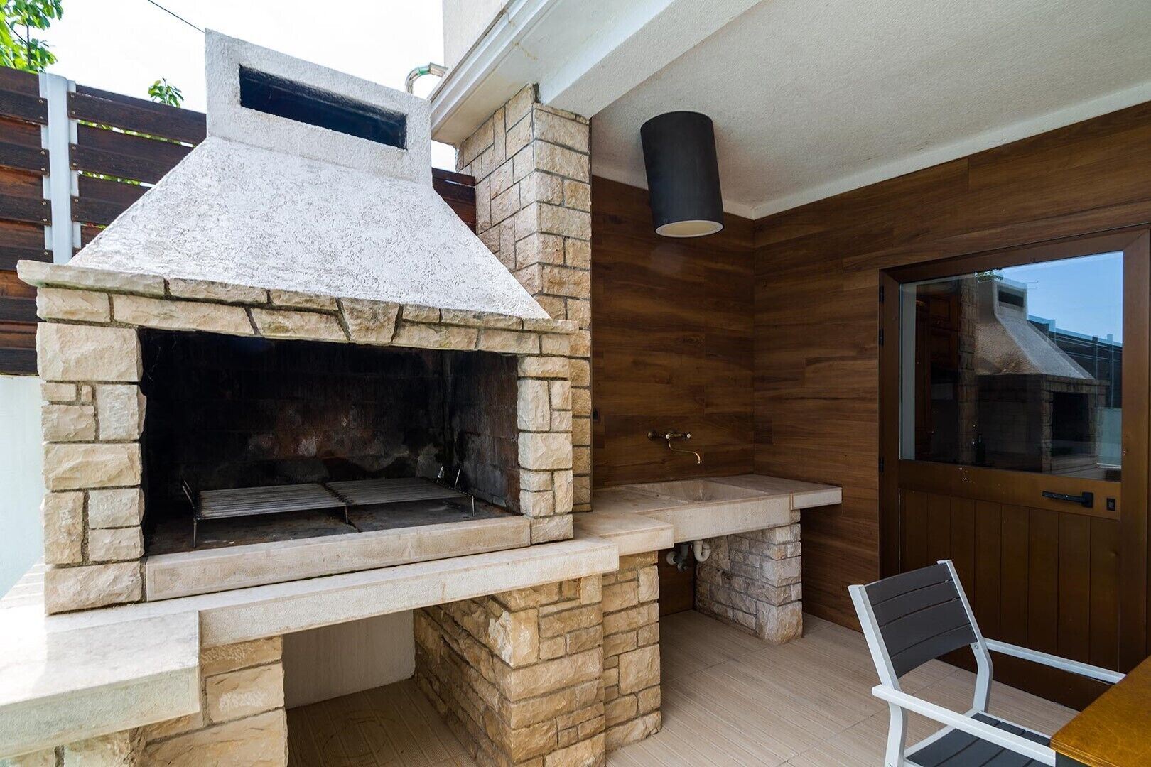 Un espace barbecue extérieur en pierre, doté d’un évier intégré et d’une chaise.
