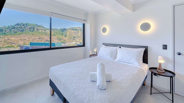 Una habitación de hotel moderna con una cama grande, dos mesitas de noche con lámparas, vista a un paisaje montañoso a través de un ventanal amplio y una lámpara de pared circular.