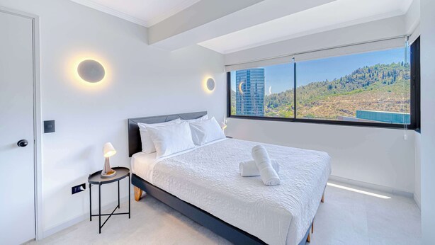 A modern bedroom with a large bed, a bedside table with a lamp, and a view of a cityscape through a large window.