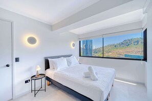 Un dormitorio moderno con una cama grande, una mesita de noche con lámpara y vistas a un paisaje urbano a través de un ventanal.