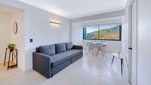 A modern living room with a grey sofa, a small table, and a dining area with chairs. A window offers a view of a landscape.