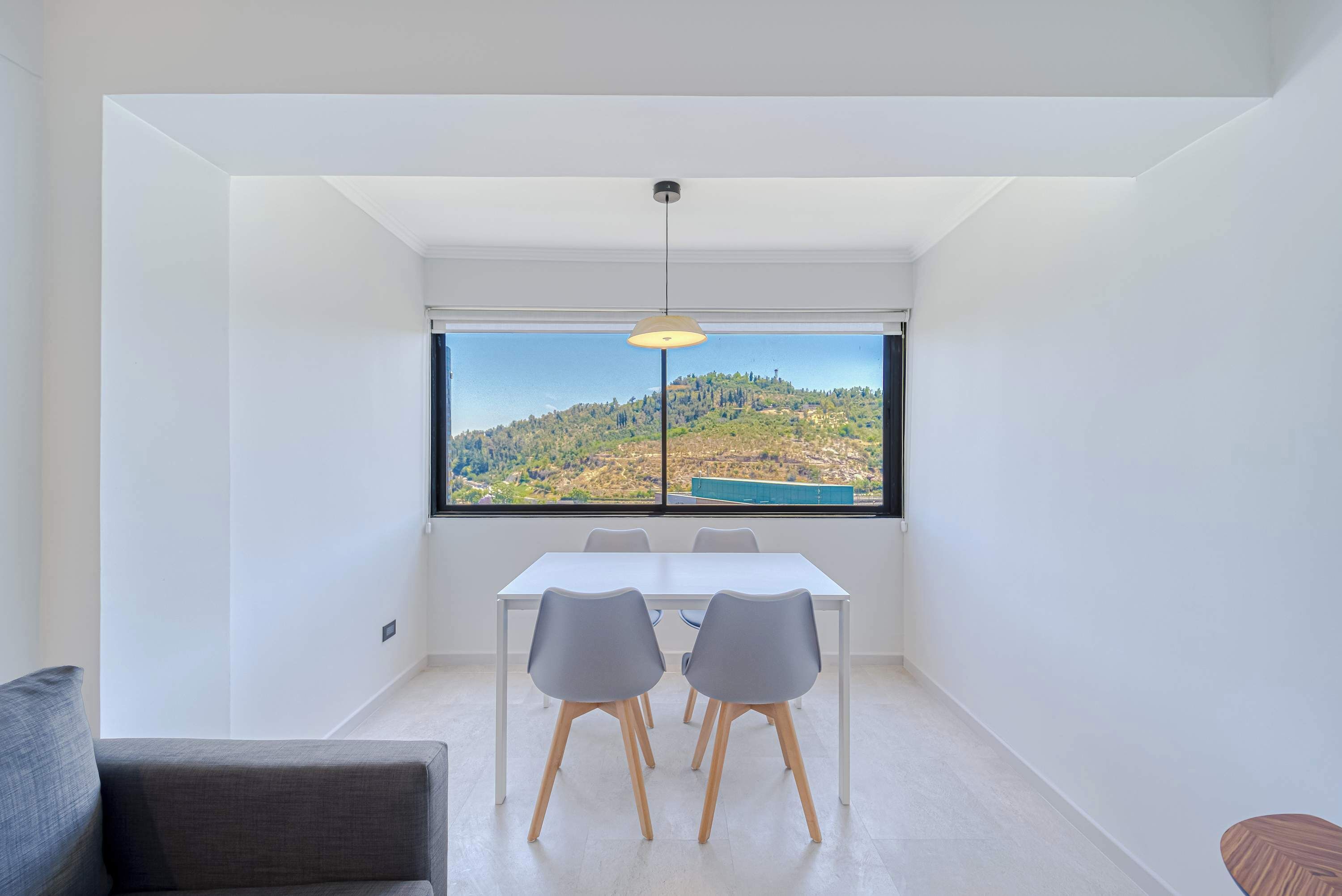 A modern room with a white table, two chairs, a sofa, and a large window with a view of a hill.