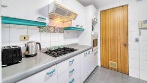Une cuisine équipée d’une cuisinière à gaz, d’une bouilloire et d’un grille-pain. Les meubles de rangement sont blancs, avec des poignées turquoise, et la porte est en bois.