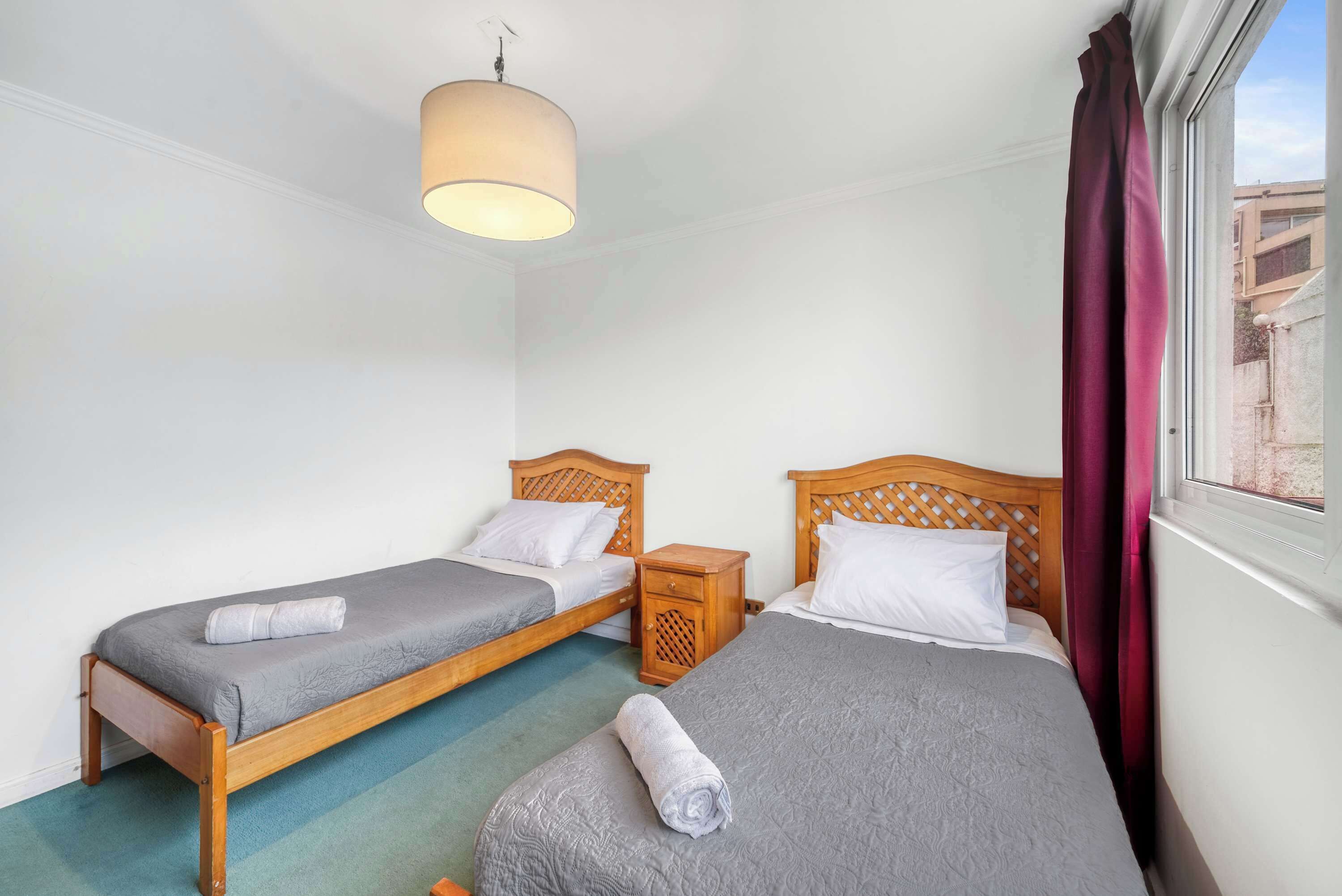 Habitación de hotel con dos camas individuales, una mesita de noche de madera, una lámpara colgante y una ventana con cortinas.