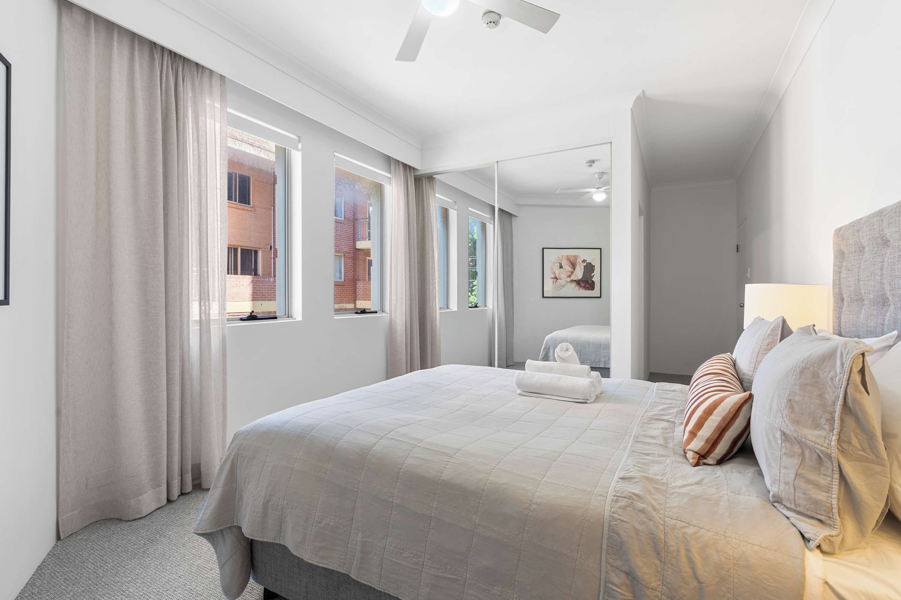 A modern hotel room with a large bed, a bathroom with a mirror, and a view of a brick building through the windows.