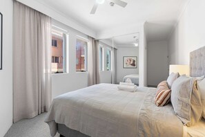 A modern hotel room with a large bed, a bathroom with a mirror, and a view of a brick building through the windows. - Camperdown Cozy Corner 1 (Camperdown)
