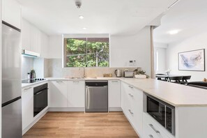 A modern kitchen with white cabinetry, a dishwasher, a built-in oven, and a window providing a view of greenery. - Camperdown Cozy Corner 1 (Camperdown)