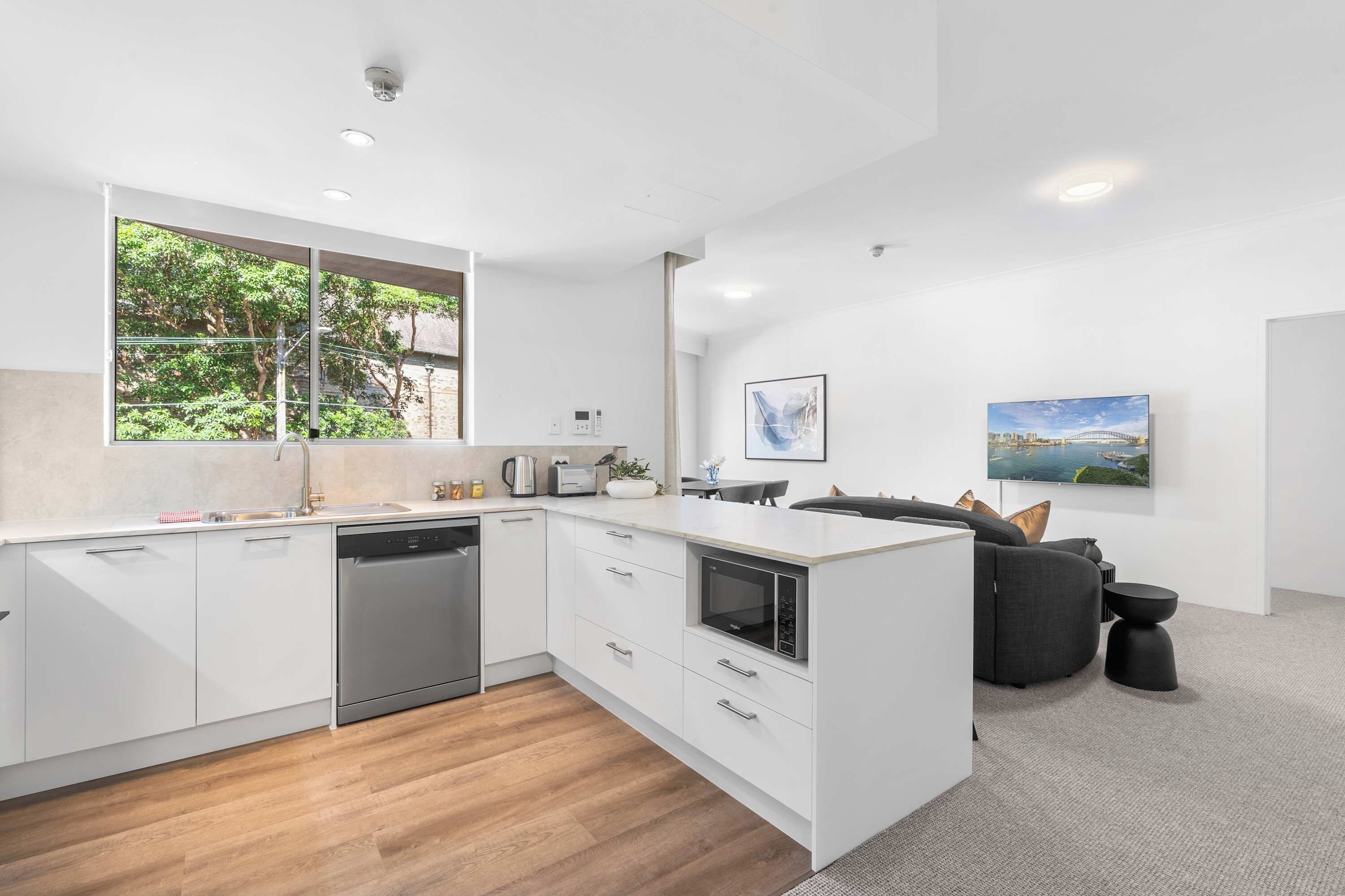 A modern kitchen with white cabinetry, a dishwasher, and a large window overlooking greenery.