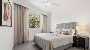 A neatly arranged bedroom with a large bed, a bedside table with a lamp, and a window with curtains.