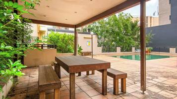 An outdoor patio with wooden tables and benches, surrounded by greenery and a swimming pool.