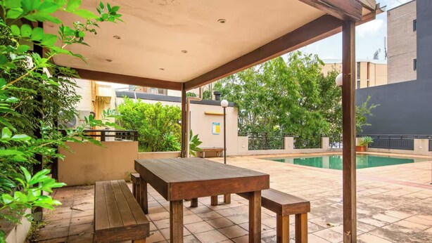 An outdoor patio with wooden tables and benches, surrounded by greenery and a swimming pool.