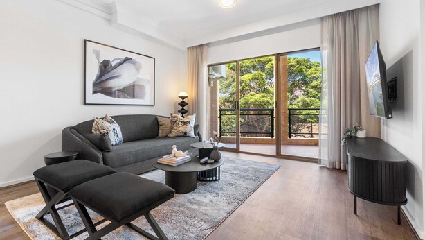 A modern living room with a grey sofa, black ottoman, and a round coffee table. There is a flat-screen TV mounted on the wall and a large window with a view of greenery outside.