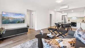 A modern living room with a flat-screen TV, a dark console table, a sofa with patterned cushions, and a dining area with a round table and chairs.