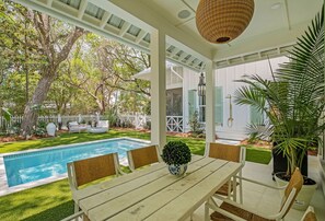 A poolside area with a table and chairs, surrounded by lush greenery and a white fence.