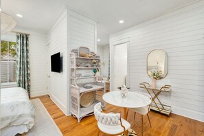 A bedroom with a bed, a white wicker storage unit, a round white table with a chair, and a wall-mounted TV.