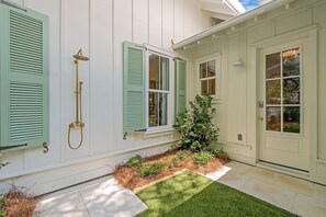 A house with white exterior walls, green shutters, a small garden with a tree, and a tiled patio.