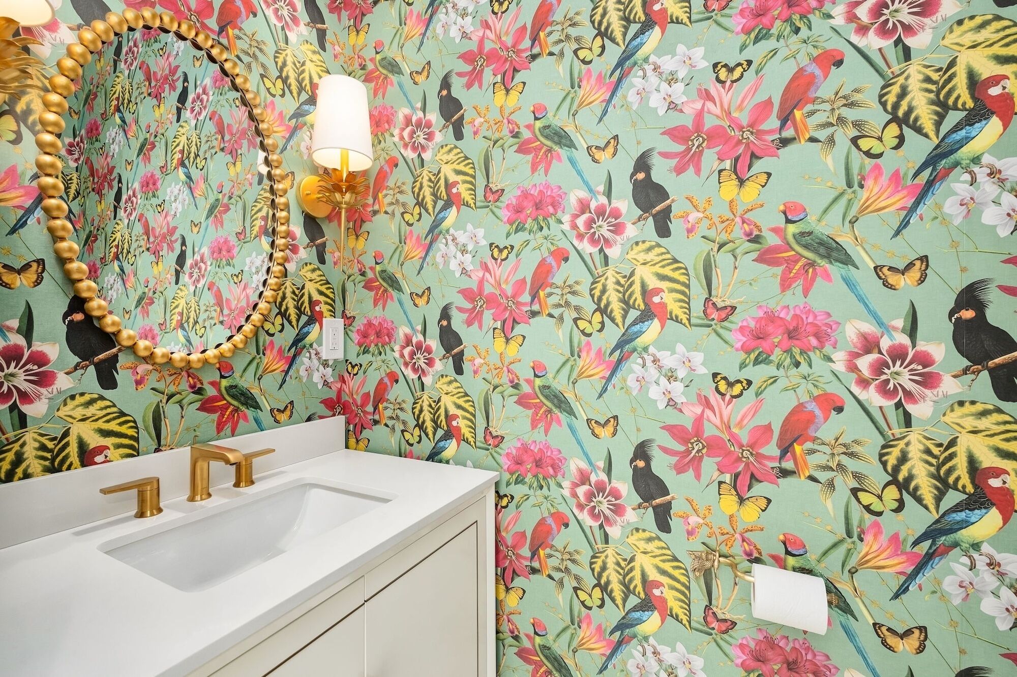 Un baño con papel tapiz floral, un collar de cuentas doradas, un lavamanos blanco y un grifo dorado.