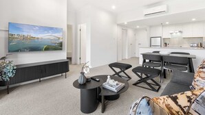 A modern living room with a large TV, a sofa, a coffee table, and a dining area with a kitchen in the background. - Camperdown Cozy Corner 8 (Camperdown)