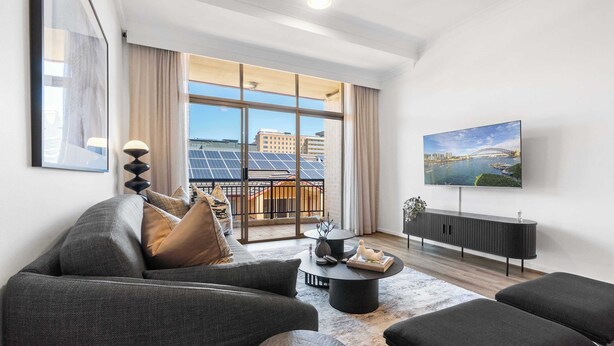 A modern living room with a sofa, coffee table, and ottoman, a TV, and a large window with a cityscape view. - Camperdown Cozy Corner 9 (Camperdown)